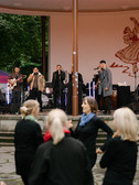 Eine Band spielt auf einer Außenbühne mit Mikrofonen, Gitarre und Saxophon. Mehrere schwarz gekleidete Personen stehen im Vordergrund und schauen zu. Im Hintergrund sind Bäume und ein großes Wandgemälde zu sehen.
