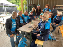 Eine Gruppe lächelnder Freiwilliger in passenden blauen Hemden sitzt und steht um einen hölzernen Picknicktisch im Freien und genießt eine Pause. Einige halten Getränke in der Hand, und im Hintergrund sind Bäume und Zelte zu sehen.