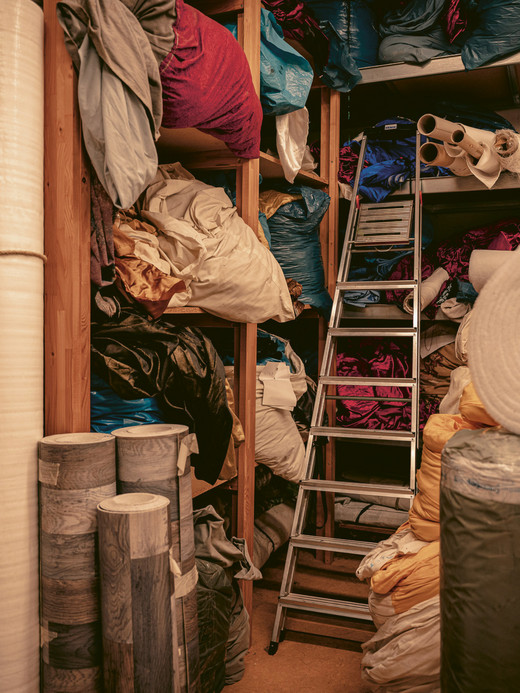 Ein Lagerraum voller Stoffrollen und verschiedener Textilien, die auf Regalen und dem Boden gestapelt sind. In der Mitte steht eine Leiter, um an die höheren Regale zu gelangen. Der Raum ist vollgestopft mit Materialien in verschiedenen Farben und Texturen.