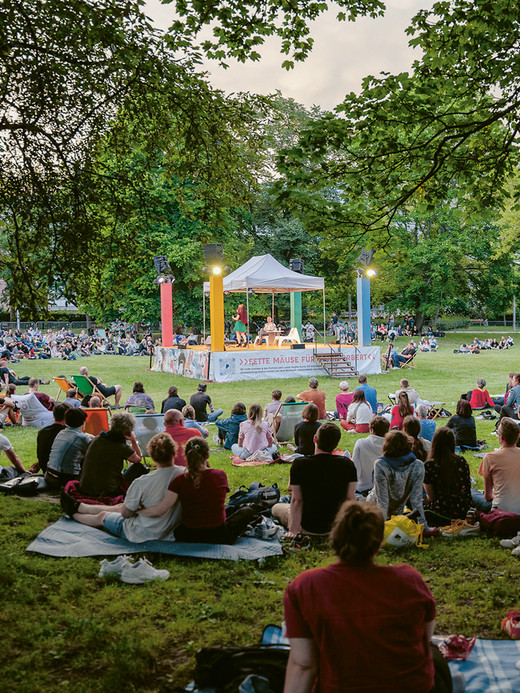 A large group of people sits on the grass in a park, watching a performance on a small, elevated stage surrounded by trees. The audience is spread out on blankets, enjoying the outdoor event under a clear sky.