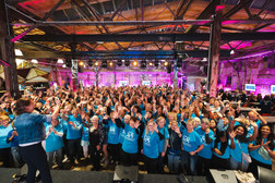 Eine große Gruppe von Menschen, viele in passenden blauen Hemden, steht in einer dekorierten Industriehalle und lächelt und winkt in die Kamera, während eine Frau auf der Bühne sie anführt. Helle Veranstaltungsbeleuchtung und festliche Atmosphäre erfüllen den Raum.