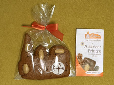 Ein Lebkuchen mit Mandeln in Form einer Hand, eingewickelt in durchsichtiges Plastik mit einer roten Schleife, neben einem Faltblatt mit der Aufschrift "Aachener Printen" auf gelbem Hintergrund.