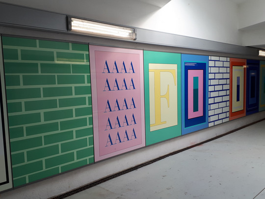 Eine Flurwand ist mit bunten Paneelen dekoriert, darunter grün-weiße Ziegelmuster, wiederholte „AAAA“-Buchstaben in Pink und große stilisierte Buchstaben mit der Aufschrift „FOOD“ in Gelb, Rot, Blau und Orange.