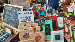 Eine bunte Auslage mit verschiedenen Schokoladentafeln und -verpackungen an einem Marktstand; auf einer Kreidetafel steht "Rette mich! Ich bin Schokolade, 5€", und ein Schild wirbt mit einem QR-Code für eine Festival-App.