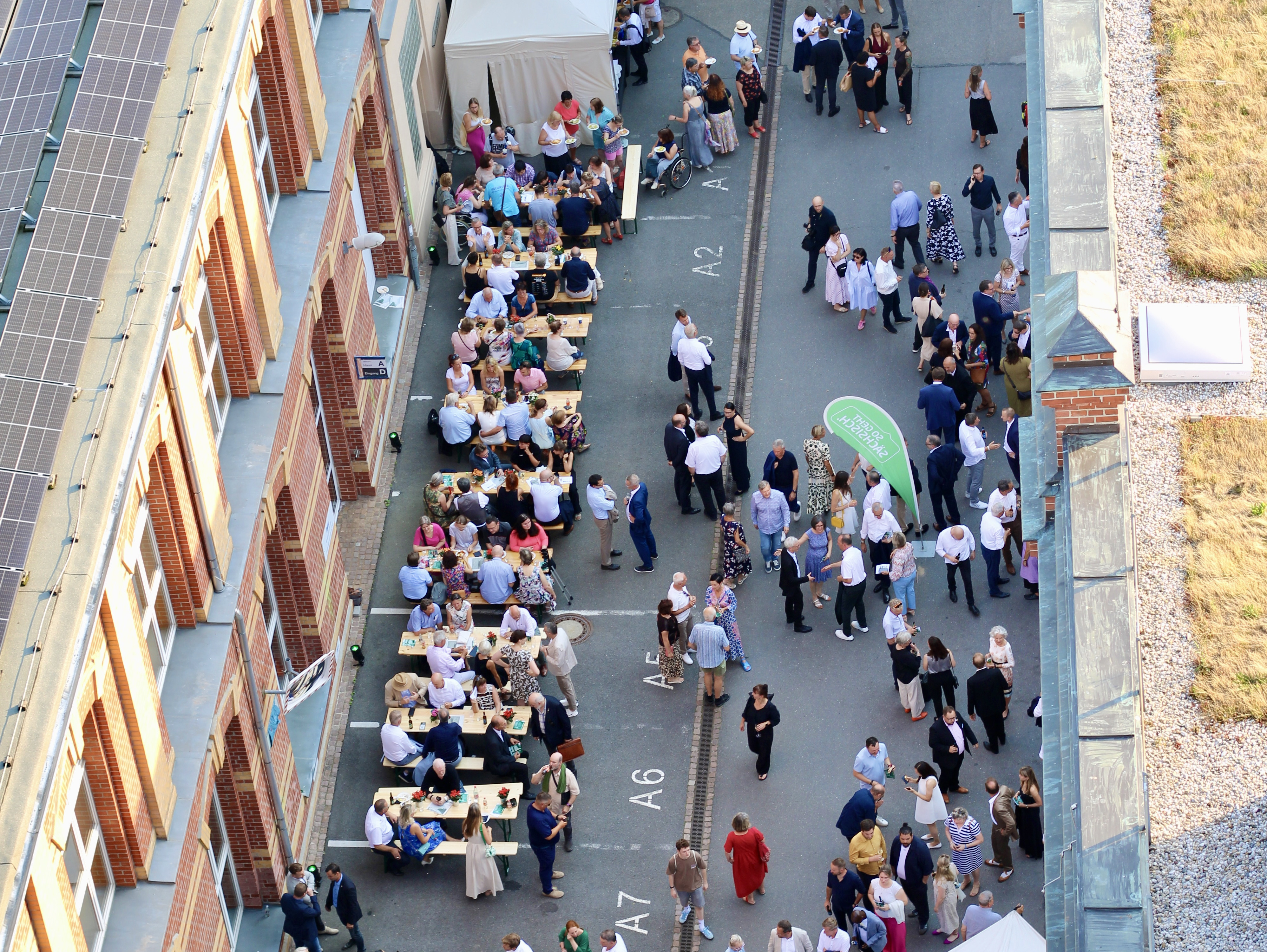 Blick von oben auf eine überfüllte Veranstaltung in einem Innenhof mit Menschen, die sich um Tische und weiße Zelte versammelt haben, neben einem Backsteingebäude mit Sonnenkollektoren auf dem Dach.