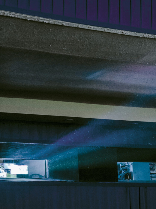 A dimly lit room with a blue and purple light beam projecting through a rectangular window. The light emits a soft glow against the dark wall, creating a misty effect. The ceiling curves overhead with an industrial feel.