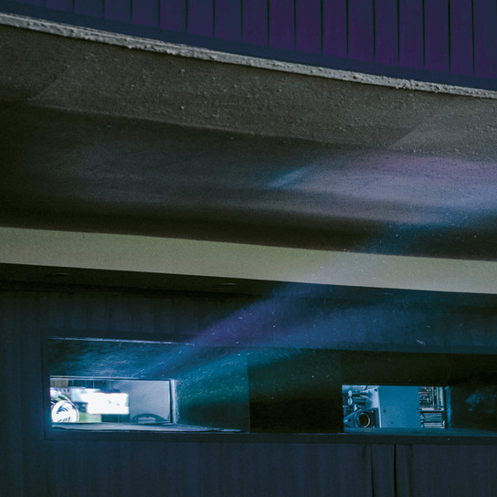 A dimly lit room with a blue and purple light beam projecting through a rectangular window. The light emits a soft glow against the dark wall, creating a misty effect. The ceiling curves overhead with an industrial feel.