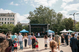 Eine Menschenmenge versammelt sich im Freien vor einer Bühne mit Künstlern. Blaue Zelte flankieren die Bühne, und im Hintergrund sind Bäume und Gebäude unter einem teilweise bewölkten Himmel zu sehen.