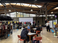 Die Menschen sitzen an Tischen in einer großen Industriehalle und beobachten eine Gruppe von Moderatoren, die auf der Bühne vor einem Bildschirm stehen. Bunte Luftballons und Dekorationen sind im Raum verstreut.