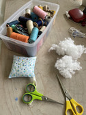 Auf einer Holzoberfläche sind eine Plastikbox mit verschiedenen Garnrollen, ein Nadelkissen mit Blumenmotiv und Stecknadeln, zwei Scheren (eine davon mit grünem Griff) und Klumpen weißer Füllwatte angeordnet.
