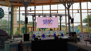 Eine hell erleuchtete Innenbühne mit einer großen Leinwand, auf der in bunten Farben "Spielzeugmacher Festival" steht. Vor der Bühne sind Kinder und Erwachsene versammelt, und durch große Fenster im Hintergrund ist das Grün im Freien zu sehen.