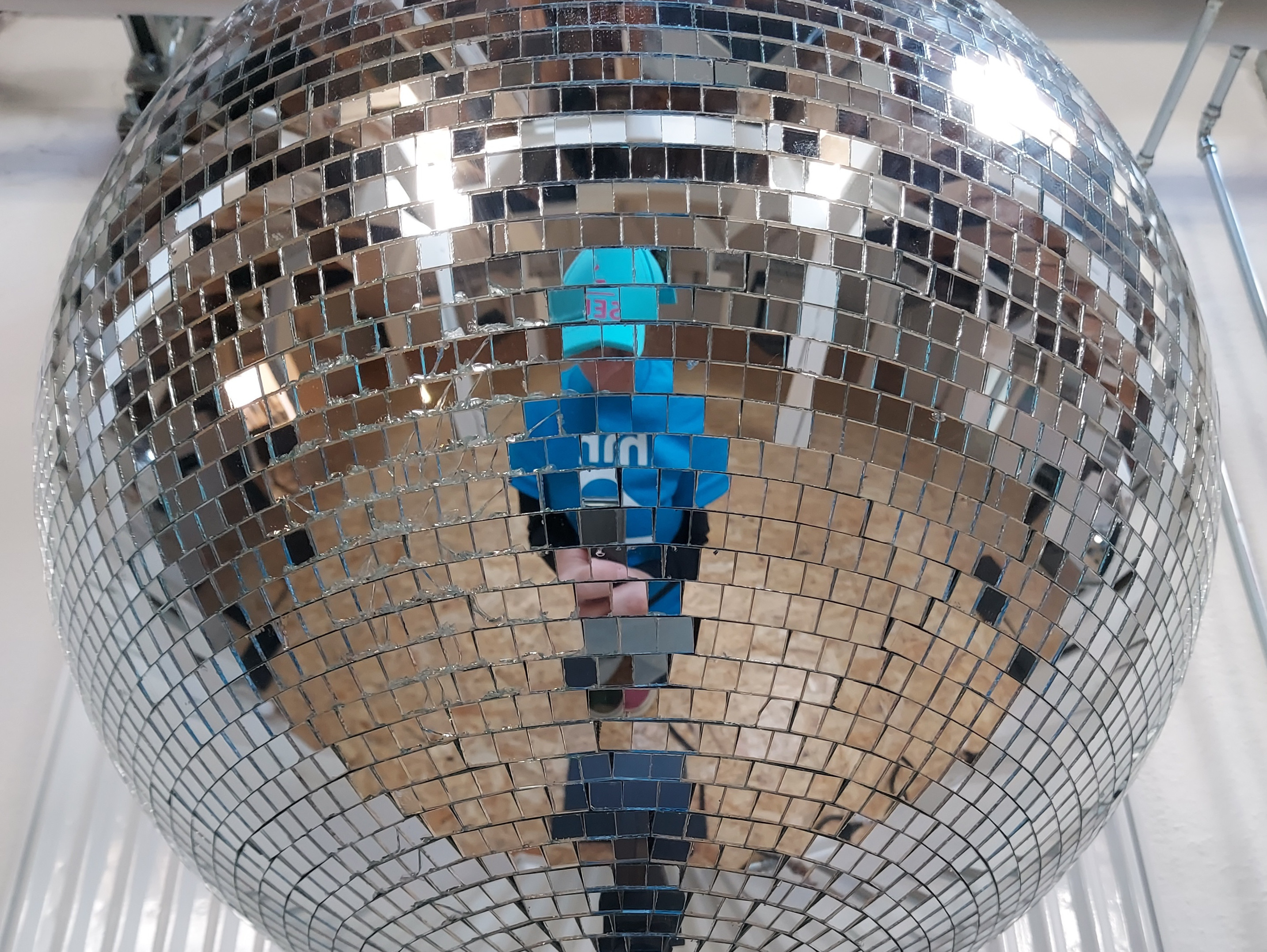 Eine Person in blauem Hemd und Hut spiegelt sich in einer großen, glänzenden Discokugel aus verspiegelten Kacheln, im Hintergrund sind ein Holzboden und Rohre zu sehen.
