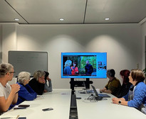 Eine Gruppe von acht Personen sitzt um einen Konferenztisch und sieht sich eine Präsentation auf einem großen Bildschirm an. Der Bildschirm zeigt drei Personen, die in einem Wald stehen. Auf dem Tisch liegen Laptops und Kabel, im Hintergrund ist ein Whiteboard zu sehen.