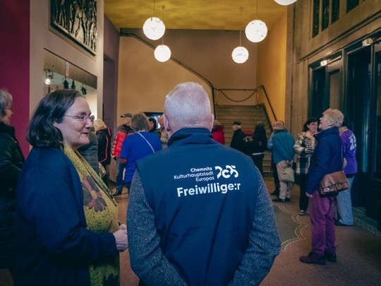 Eine Gruppe von Menschen steht in der Eingangshalle eines Gebäudes und unterhält sich. Eine Person trägt eine Weste mit der Aufschrift „Freiwillige:r“. Die Atmosphäre ist entspannt; andere sind versammelt oder gehen auf eine Treppe im Hintergrund zu. Kugelförmige Lichter hängen von der Decke.