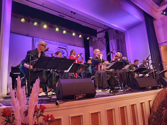 Eine Band aus Musikern und Sängern tritt auf einer Bühne mit Mikrofonen und Notenständern auf, die von bunten Bühnenlichtern in einem großen Innenraum mit violetter Beleuchtung angestrahlt wird.