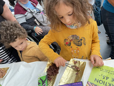 Ein junges Mädchen in einem gelben Hemd schneidet Schokoladenstücke auf einem goldenen Tablett, während ein kleiner Junge neben ihr an einem mit Papieren und Schokoladenverpackungen bedeckten Tisch zusieht.