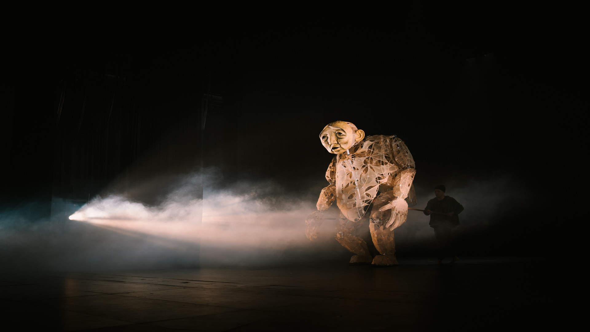 Eine große, menschenähnliche Puppe mit traurigem Gesichtsausdruck wird von einem Lichtstrahl auf der Bühne angestrahlt und von Nebel umgeben, während eine Person im dunklen Theater teilweise dahinter steht.