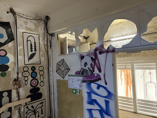 Ein Raum mit abblätternder Farbe, geometrischer Wandgestaltung und Graffiti mit einem Panda auf einem lila Autoscooter. Es gibt freiliegende Drähte, Notausgangsschilder und ein Fenster mit ausgeschnittenen Bögen über der Tür.