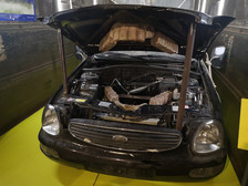 Ein schwarzer Ford mit geöffneter Motorhaube zeigt einen ungewöhnlichen Motorraum, der anstelle eines herkömmlichen Motors mit Ziegelsteinen gefüllt ist und auf einem gelben Boden und Metallwänden steht.