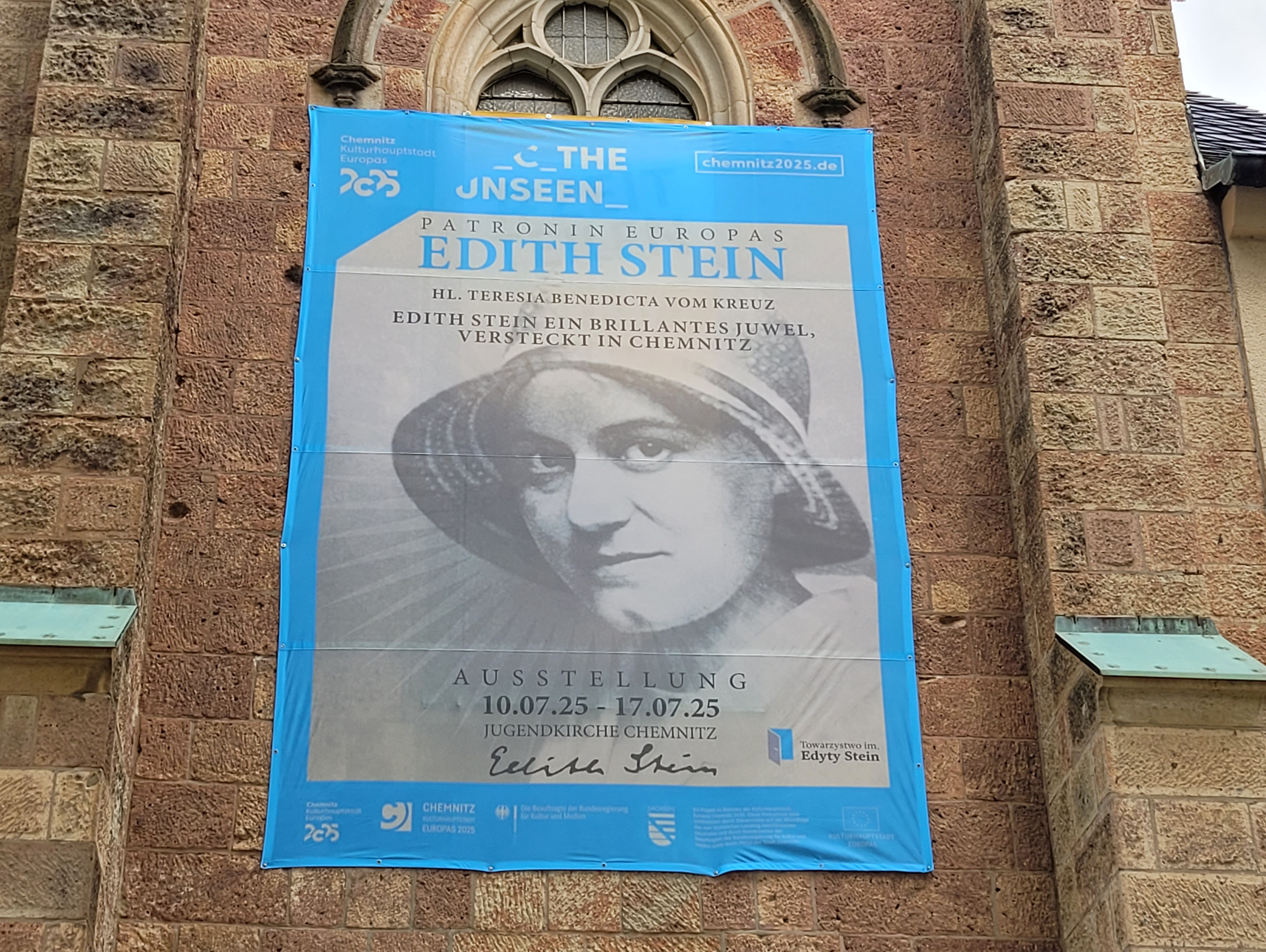 Auf einem blauen Banner an einer gemauerten Kirchenwand ist ein schwarz-weißes Porträt von Edith Stein mit Hut abgebildet. Der Text kündigt eine Ausstellung über sie mit dem Titel "Das Unsichtbare" in Chemnitz, Deutschland, vom 10. Juli 2025 bis zum 17. Juli 2025 an.