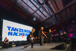In einem geräumigen Saal mit freiliegenden Holzbalken und einem auf beiden Seiten sitzenden Publikum führt eine Gruppe von Personen auf der Bühne einen Tanz vor einer großen Leinwand auf, auf der die Worte "Tanzende Nachbarn" zu lesen sind.