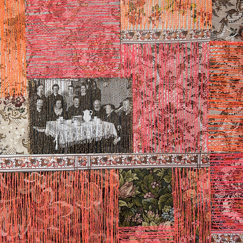[Translate to Englisch:] Ein Patchwork aus floralen und gemusterten Stoffquadraten, hauptsächlich in Rosa- und Orangetönen, mit vertikal herabhängenden orangefarbenen Fäden. Ein Quadrat enthält ein Schwarzweißfoto von Personen, die an einem Tisch sitzen.
