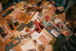 Eine Gruppe von Menschen sitzt um einen Tisch herum, der mit buntem Papier, Markern, Stiften, Karten und Bastelmaterial bedeckt ist, und beschäftigt sich mit Bastelarbeiten.