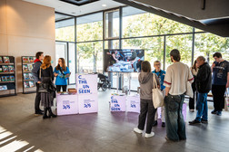 Menschen versammeln sich in einem hellen Foyer an einem Veranstaltungsstand mit beleuchteten Blöcken, auf denen "THE UNSEEN" steht. Freiwillige Helfer in blauen Hemden unterhalten sich mit den Besuchern, während im Hintergrund ein großer Bildschirm eine Ausstellung zeigt.