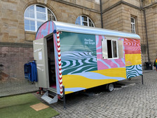 Ein bunter Wohnwagen mit der Aufschrift "Pavillon der Angst" steht auf einer Kopfsteinpflasterstraße vor einem Gebäude mit Bogenfenstern und Steinmauern. Der Anhänger ist mit wellenförmigen, mehrfarbigen Mustern versehen.