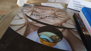 Nahaufnahme eines Tisches, auf dem das Buch "Rebecca Horn: Glowing Core" mit einem architektonischen Titelbild. In der Nähe befinden sich gestapelte Broschüren und ein teilweise sichtbares Buch in deutscher Sprache.