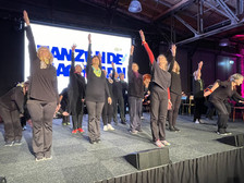 Eine Gruppe schwarz gekleideter älterer Erwachsener führt auf der Bühne einen Synchrontanz auf und streckt dabei einen Arm in die Höhe. Im Hintergrund sind ein großes beleuchtetes Schild und eine Decke im Industriestil mit sichtbaren Balken zu sehen.