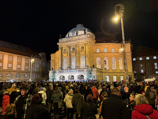 Eine große Menschenmenge versammelt sich nachts vor einem verzierten, beleuchteten historischen Gebäude mit Rundbogenfenstern. Einige Weihnachtsbäume und Lichter schmücken den Platz und schaffen eine festliche Atmosphäre. Die Menschen tragen Winterkleidung.