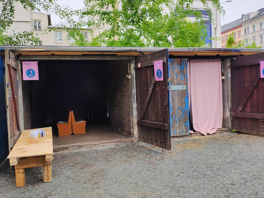 Drei Holzstände mit offenen Türen sind mit den Nummern 1, 2 und 3 gekennzeichnet. Stand 1 hat einen Tisch mit Papieren davor, Stand 2 ist leer und Stand 3 hat einen rosa Vorhang. Der Bereich ist mit Kiesboden und Gebäuden im Hintergrund bedeckt.