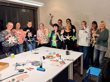 Eine Gruppe lächelnder Menschen steht in einem gut beleuchteten Raum und hält bunte Papiersterne. Sie sind um einen Tisch mit Bastelmaterial, Papier und Snacks versammelt, was auf einen kreativen Workshop oder eine Bastelveranstaltung hindeutet.