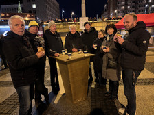 Eine Gruppe von acht Erwachsenen steht nachts im Freien um einen hohen Holztisch, hält Tassen und Essen in der Hand, während im Hintergrund eine Menschenmenge und beleuchtete Stadtgebäude zu sehen sind.