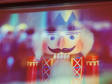 Eine Nahaufnahme einer bunten hölzernen Nussknackerfigur mit bemaltem Gesicht, schwarzem Schnurrbart, rosigen Wangen, weißem flauschigen Haar und einer Krone vor einem unscharfen, bunten Hintergrund.