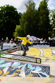 Ein junger Skateboarder in gelber Jacke und weißem Helm führt einen Trick auf einer Schiene in einem Skatepark im Freien aus, mit buntem Graffiti und einer Menge von Zuschauern im Hintergrund.