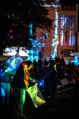 Menschen stehen nachts im Freien, halten und interagieren mit reflektierenden Tafeln, während bunte Lichter und Projektionen sie und die Umgebung, einschließlich eines Gebäudes im Hintergrund, beleuchten. Die Atmosphäre ist lebendig und künstlerisch.
