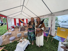 Zwei Frauen stehen lächelnd in einem weißen Zelt, das mit bunten Stoffen und spirituellen Gegenständen geschmückt ist. Sie halten Federn und tragen traditionelle Kleidung. Kissen und zeremonielle Gegenstände sind auf dem Boden um sie herum angeordnet.