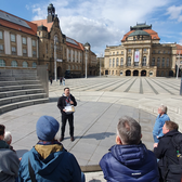 Ein Reiseleiter spricht zu einer kleinen Gruppe von Menschen auf einem offenen, sonnigen Stadtplatz, umgeben von historischen Gebäuden mit verzierten Fassaden und roten Dächern. Der Himmel ist teilweise bewölkt.