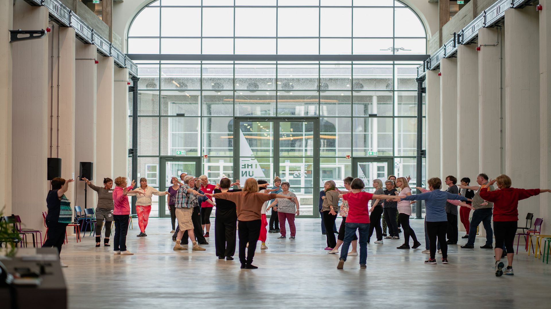 Eine Gruppe Erwachsener steht in einer geräumigen, hellen Halle mit hohen Decken und großen Fenstern und nimmt mit ausgestreckten Armen an einer Gruppenaktivität teil, wobei sie einen Kreis bilden.