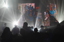 Silhouettierte Menschen stehen bei einer Indoor-Veranstaltung vor einer hellen Bühnenbeleuchtung und einem großen Bildschirm, auf dem ein Gruppenfoto zu sehen ist, was eine lebendige und dynamische Atmosphäre schafft.