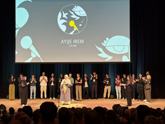 Eine Gruppe von Menschen steht auf einer Bühne, einige klatschen, während Ayşe İrem in der Mitte steht und Blumen hält. Auf einem Bildschirm dahinter sind ihr Name und "İSLAM" mit Mikrofon und abstrakten Grafiken zu sehen.