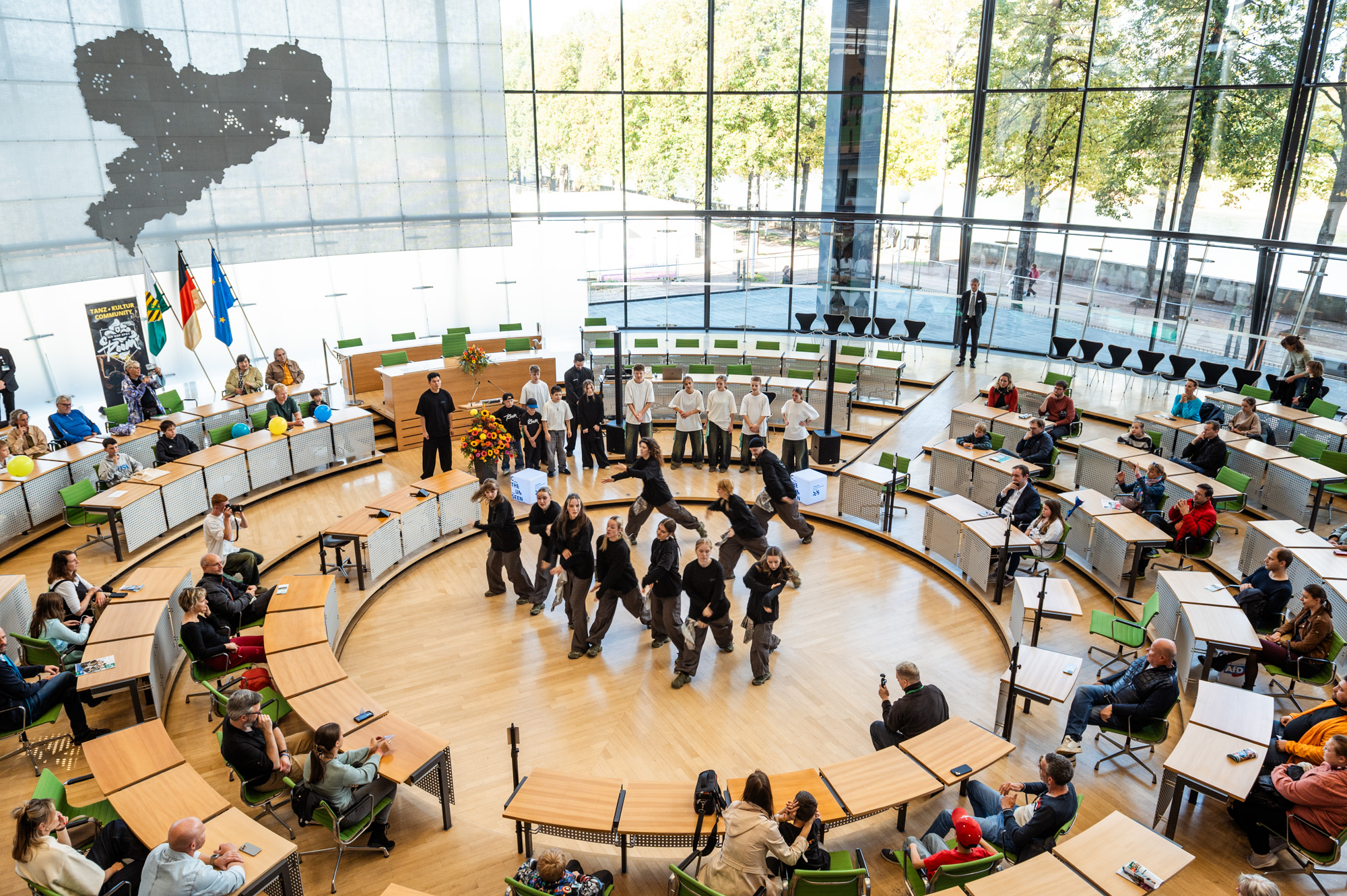 A group performs a dance in the centre of a round, modern auditorium with large windows. People sit around and watch, and flags are placed on the left. Outside there are trees and sunlight.