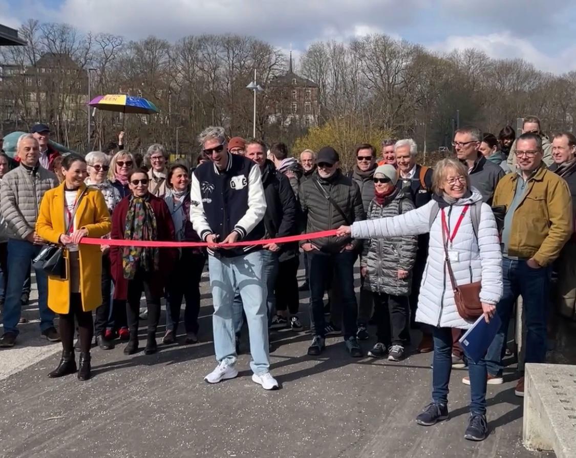 Eine Gruppe lächelnder Menschen, während ein Mann und eine Frau im Freien ein rotes Band halten und durchschneiden, um die Eröffnung eines Gehwegs oder einer Veranstaltung zu markieren. Im Hintergrund sind Bäume und Gebäude zu sehen.