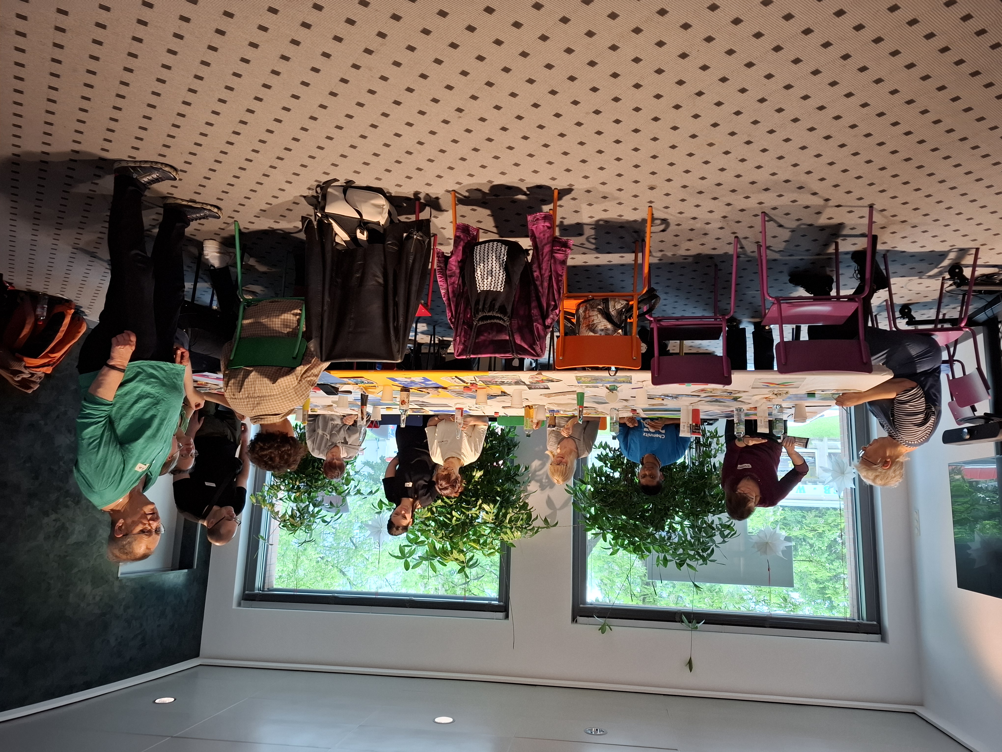 Eine Gruppe von Menschen sitzt und steht in einem hellen Raum mit großen Fenstern, Pflanzen und bunten Stühlen um einen mit Kunstbedarf bedeckten Tisch und ist mit einer kreativen Aktivität oder einem Workshop beschäftigt.