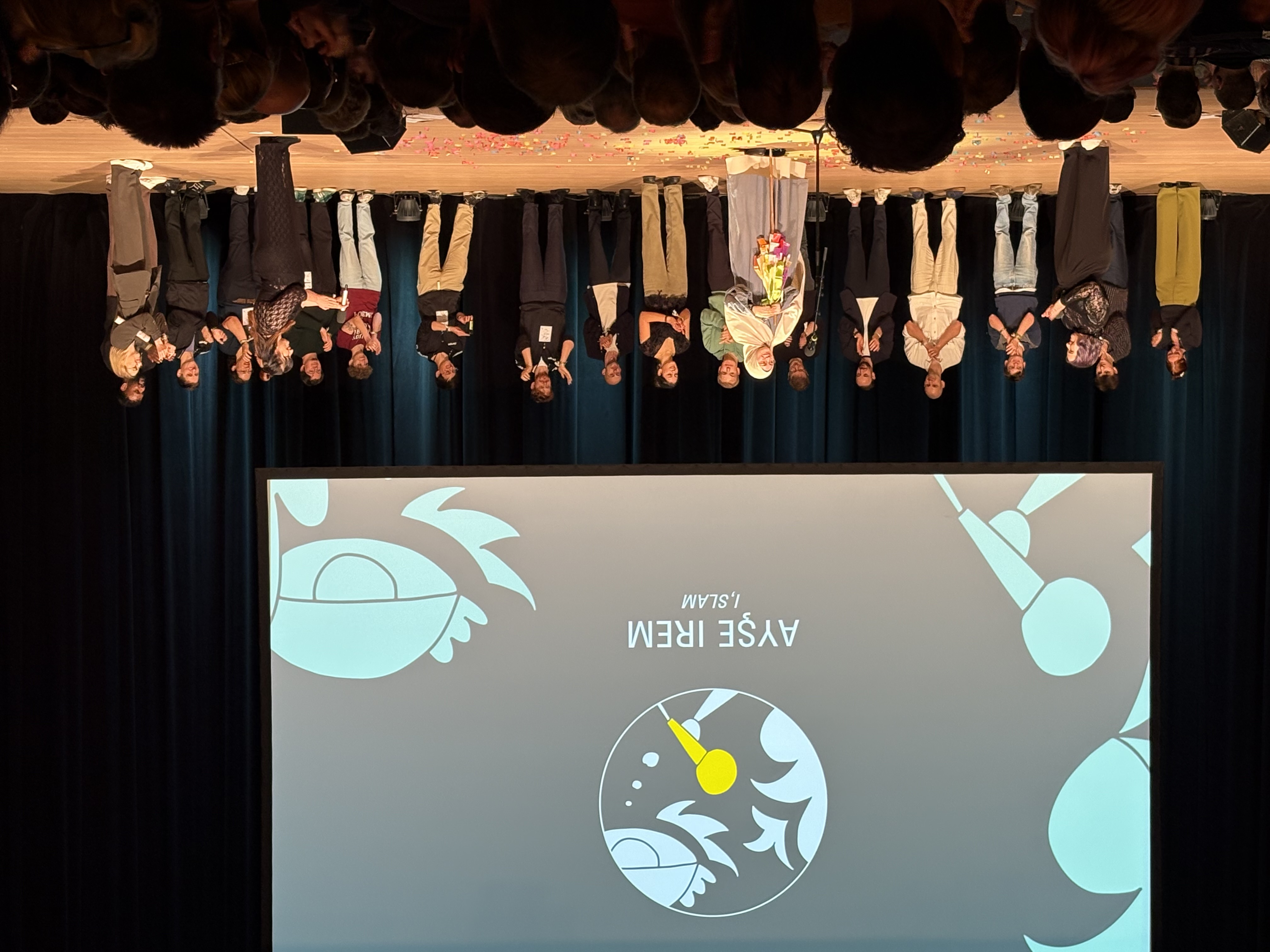 Eine Gruppe von Menschen steht auf einer Bühne, einige klatschen, während Ayşe İrem in der Mitte steht und Blumen hält. Auf einem Bildschirm dahinter sind ihr Name und "İSLAM" mit Mikrofon und abstrakten Grafiken zu sehen.