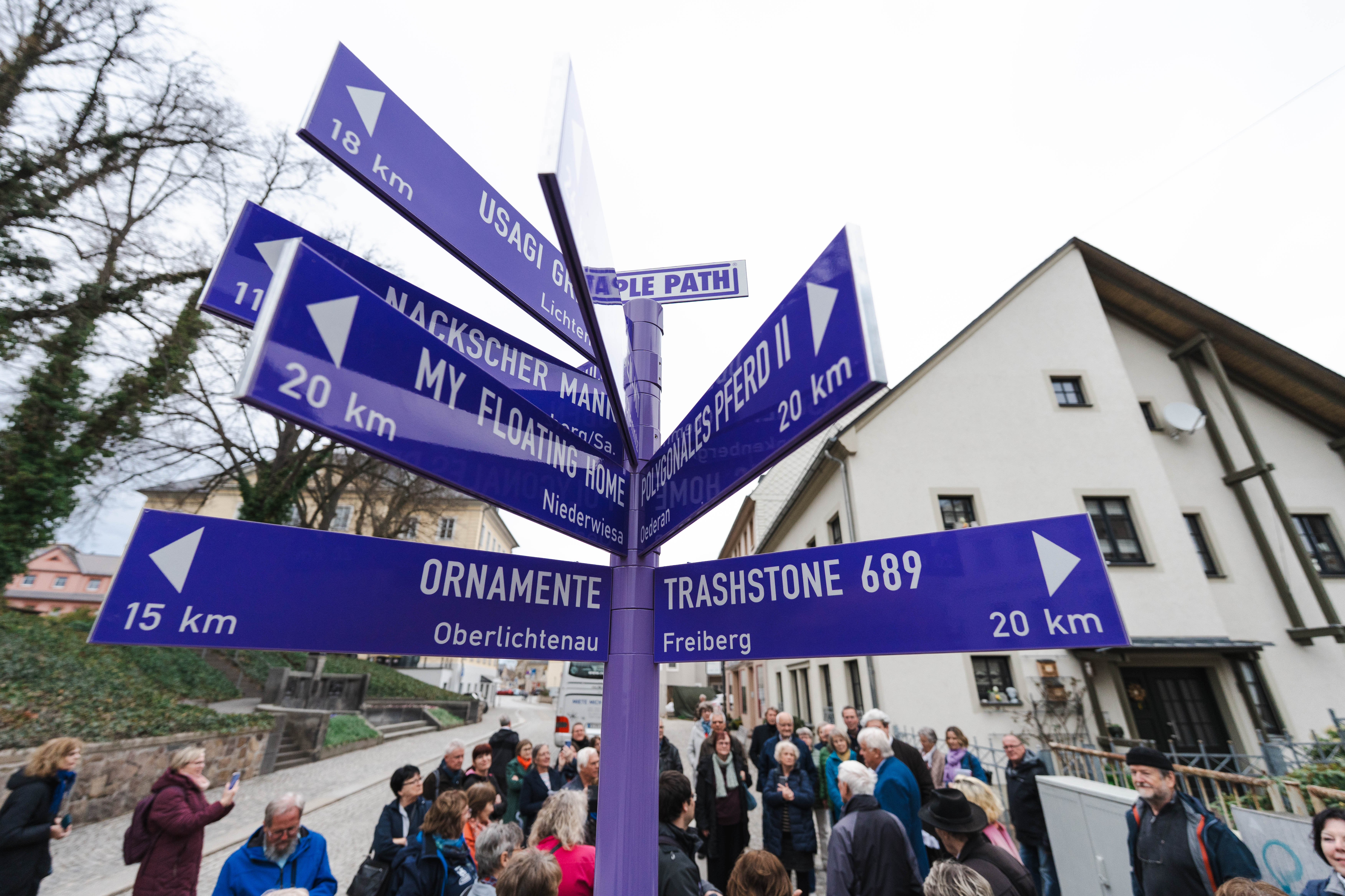 A group of people stand around a purple signpost at the PURPLE PATH in Hainichen with several arrows pointing in different directions, each labelled with destination names and distances in kilometres.